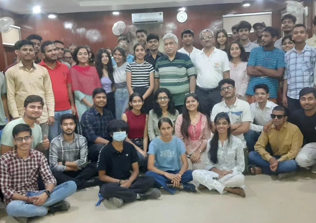 A large gathering of about 40-50 young adults, predominantly students, are posing for a group photo in a warmly lit room with wooden paneling. Some are seated cross-legged on the floor, while others stand behind them, all smiling. Two older men, possibly faculty or organizers, are also present in the center of the group.