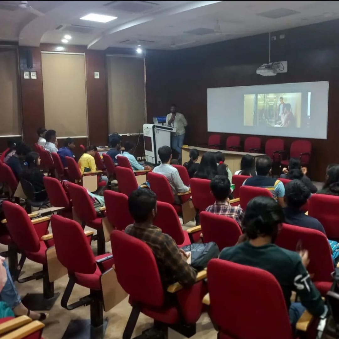 A speaker presents in a darkened lecture hall, with an image projected on a large screen. An audience of around 30 individuals is seated in rows of red chairs, facing the front.