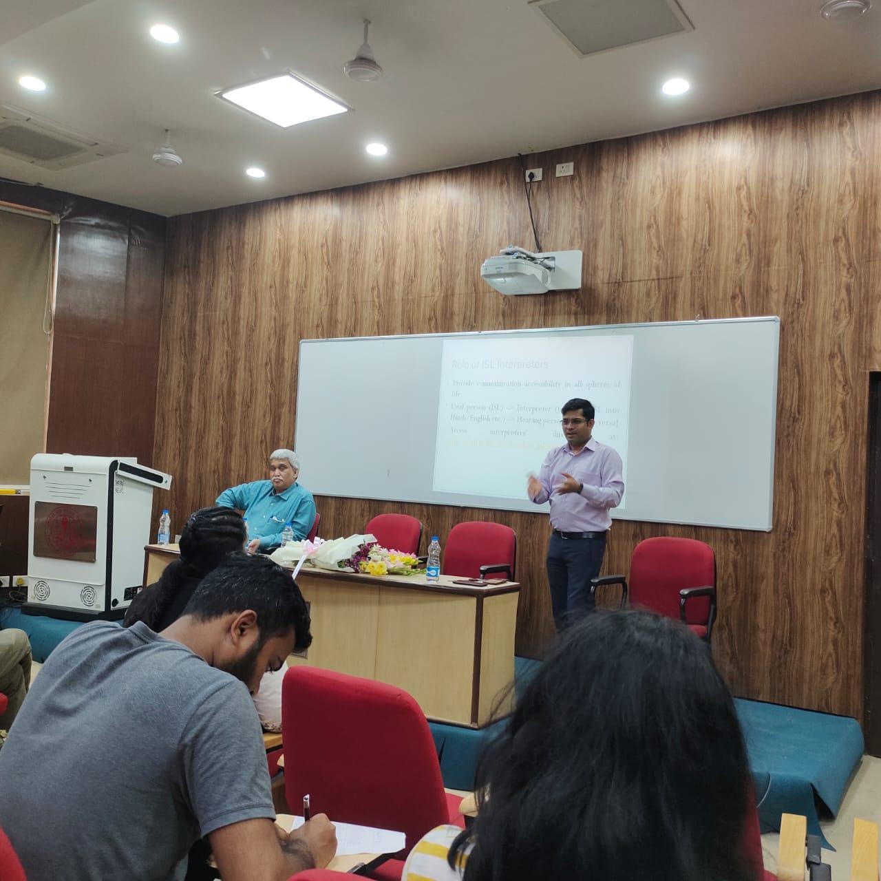 A male speaker in a light purple shirt and dark trousers gestures while presenting in front of a whiteboard to an audience in a lecture hall. Another man sits at a table with flowers.