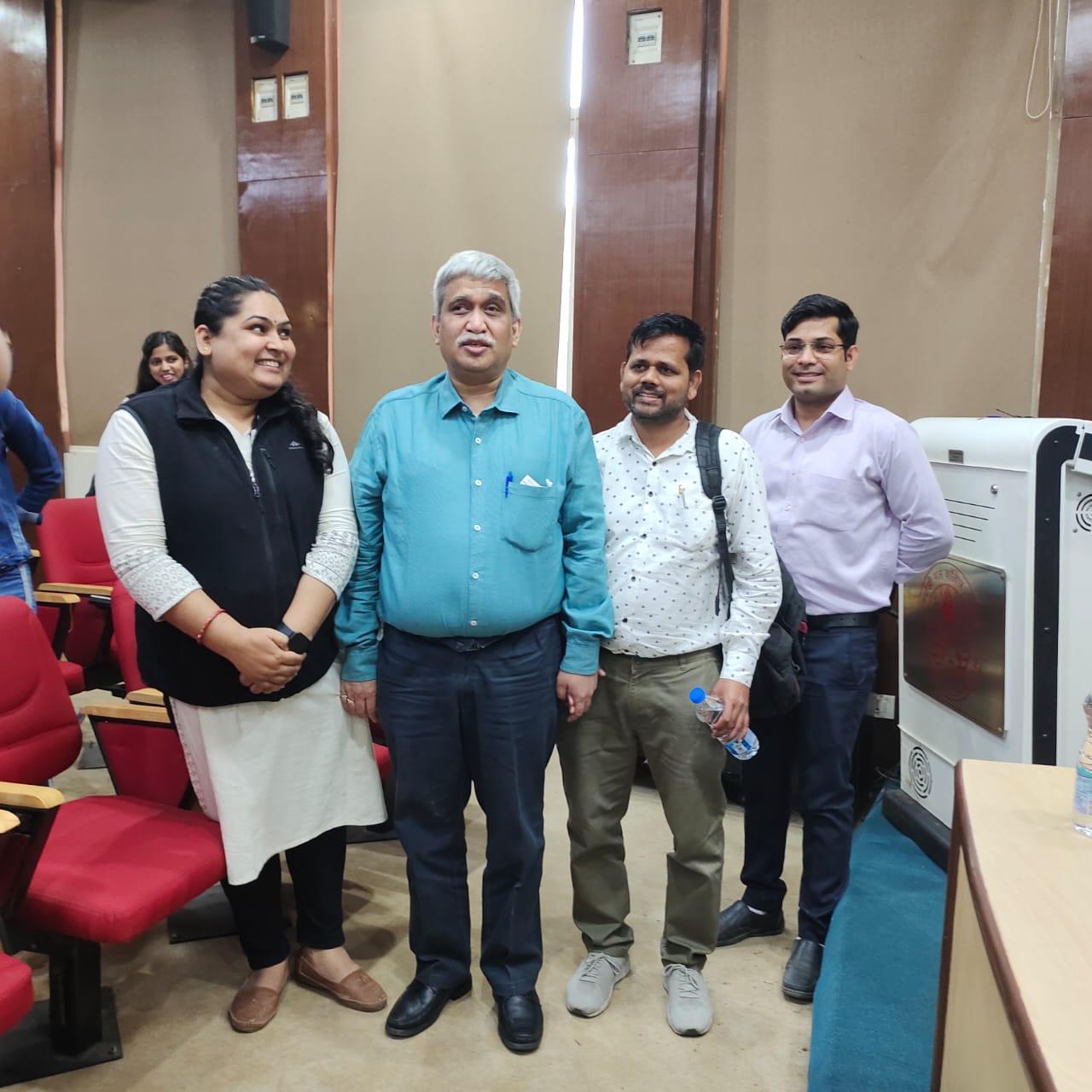 Four people stand smiling in an auditorium setting with red seats. Two men in collared shirts flank a man in a teal shirt with grey hair, and a woman in a black vest and white kurta stands to the far left.