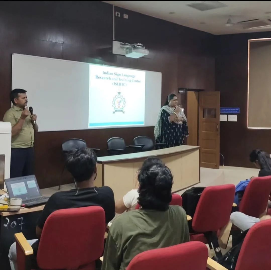 A presentation is taking place in a lecture hall. A man in a light green shirt speaks into a microphone on the left, while a woman in a dark patterned dress stands behind a podium on the right. A screen displays "Indian Sign Language Research and Training Center (ISLRTC)" and a logo. A small audience is seated, with two individuals in the foreground facing away from the camera.
