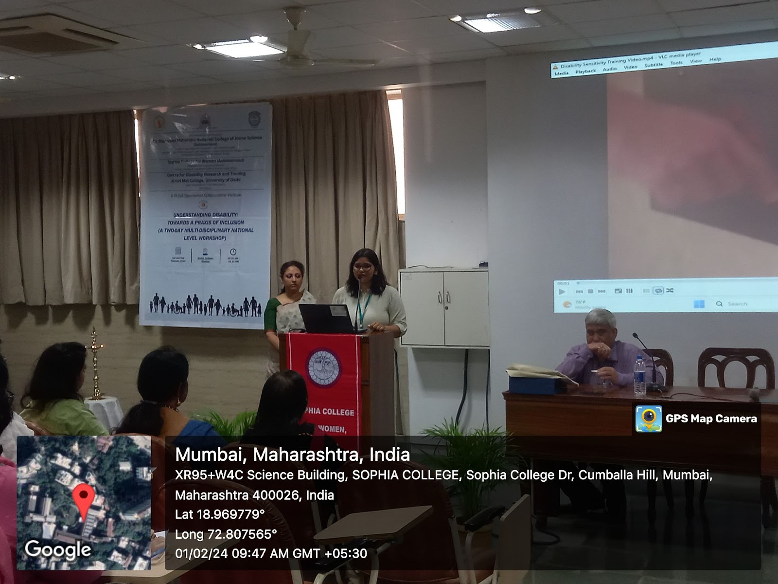 A presentation is underway in a lecture hall. Two women stand at a podium, addressing a seated audience. A projector screen displays text, and a map shows the location as Sophia College, Mumbai, India
