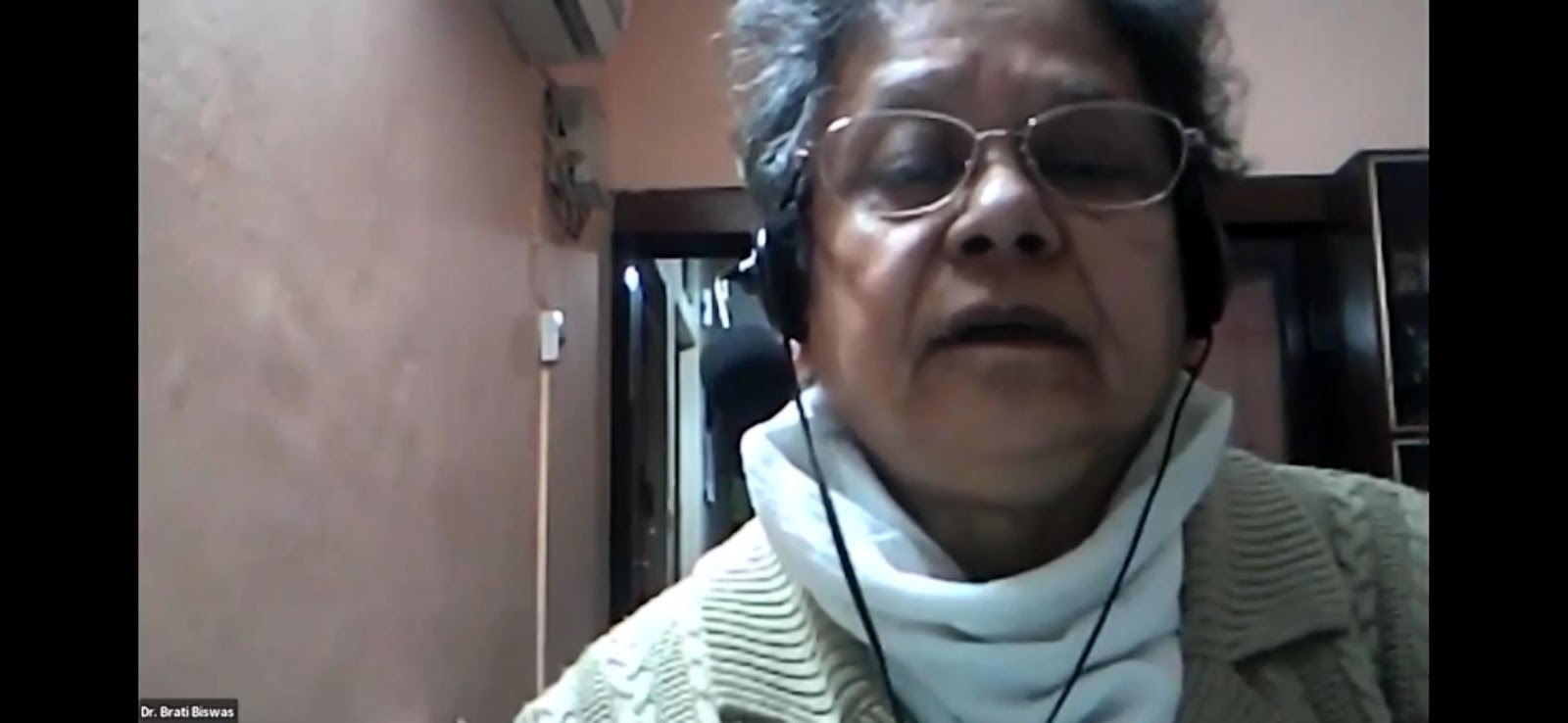 A close-up video call still of Dr. Brati Biswas, an older woman with short grey hair and glasses, wearing a light-colored sweater and a white scarf, speaking.
