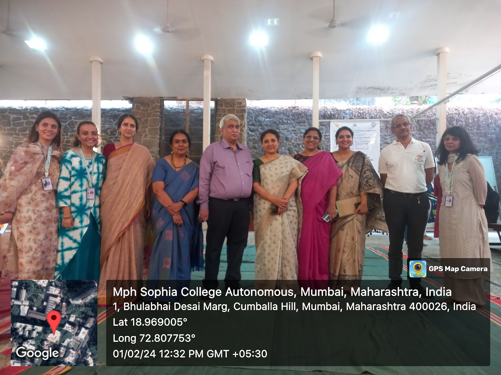 
A group of nine professionals, including several women in saris and one man in a light purple shirt, stand together for a photo in an outdoor setting with pillars.