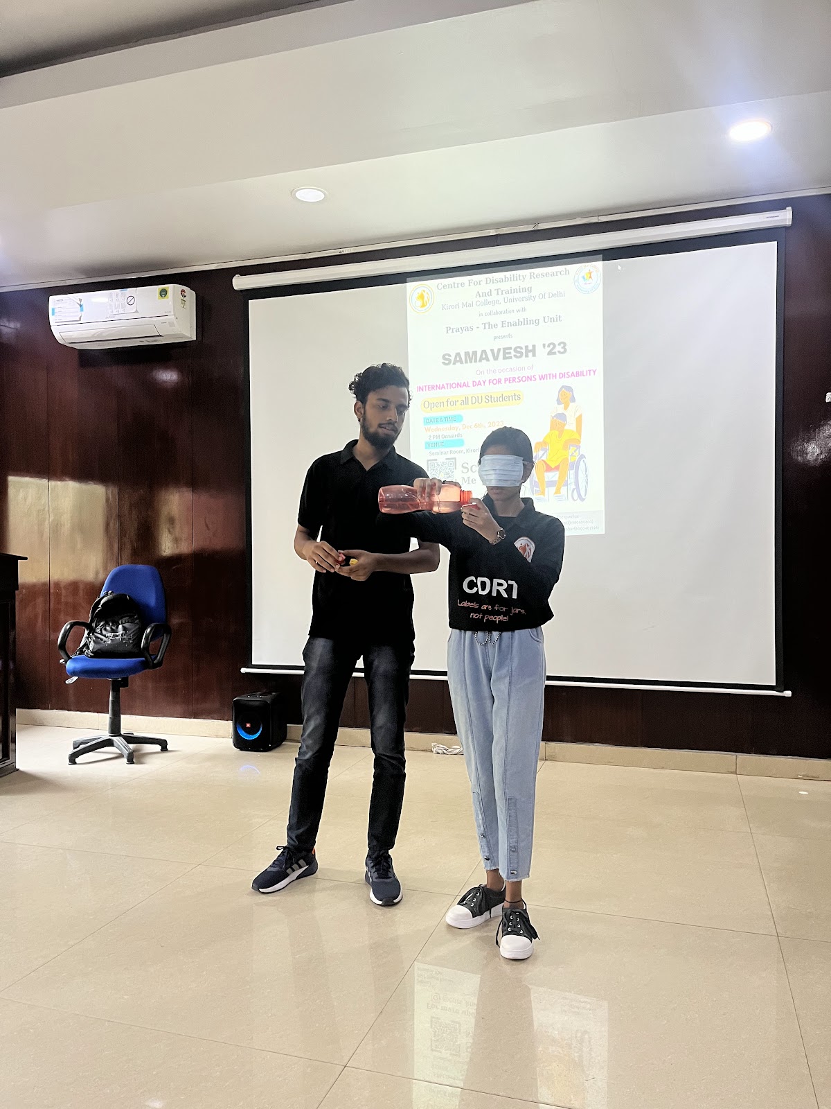 A male instructor stands next to a blindfolded student holding an object, in front of a projection screen displaying a poster for "SAMAVESH '23" on International Day for Persons with Disability.