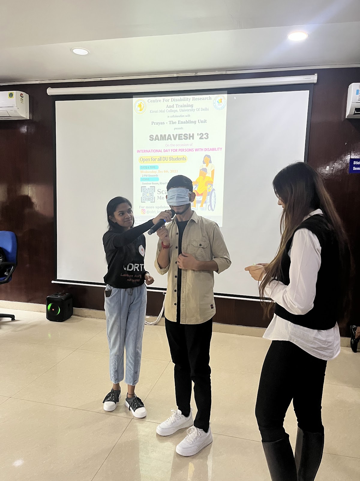 A female student holds a microphone for a blindfolded male student during a presentation, with a female instructor observing. A poster for "SAMAVESH '23" is projected behind them.