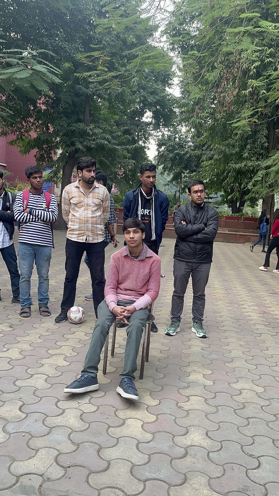 A young man in a striped sweater sits on a chair outdoors, surrounded by other men standing. They are on a paved area with trees in the background, possibly observing an activity.
