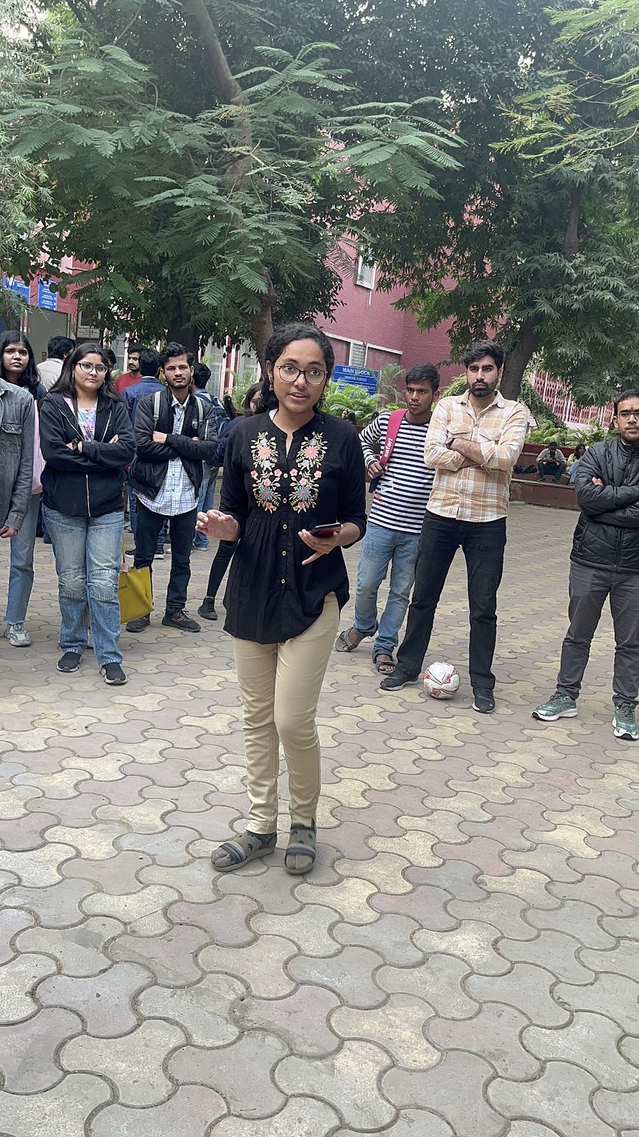 A young woman in a black embroidered top and beige pants speaks to a group outdoors about visual impairments. She stands on a paved surface with trees and buildings in the background, facing the camera.