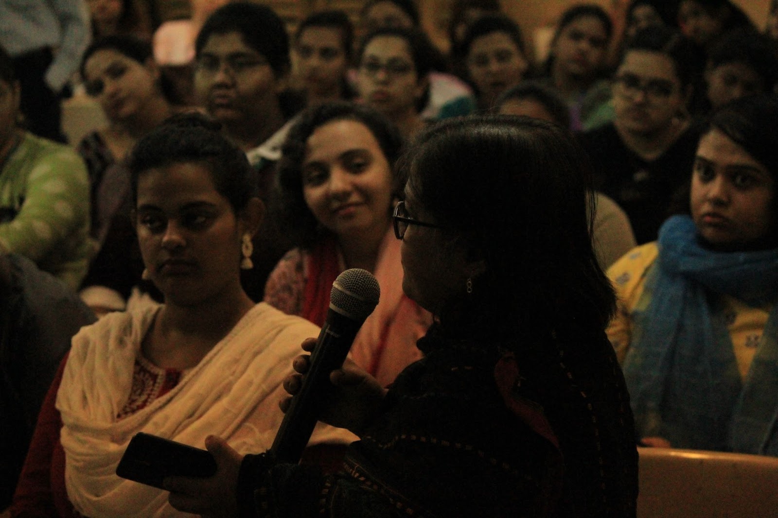 A young woman, seen from behind, holds a microphone and speaks to a seated audience, whose faces are partially visible in the softly lit room.