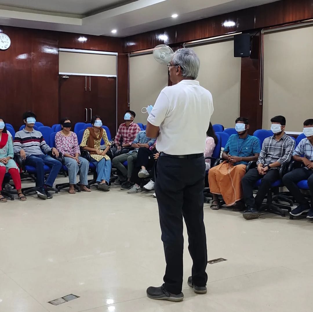 A man in a white polo shirt stands facing a semi-circle of about 15-20 individuals seated in blue chairs. Most of the seated participants are wearing blindfolds or eye masks, suggesting an interactive sensory workshop in progress. The room has wooden paneling and a projector screen.