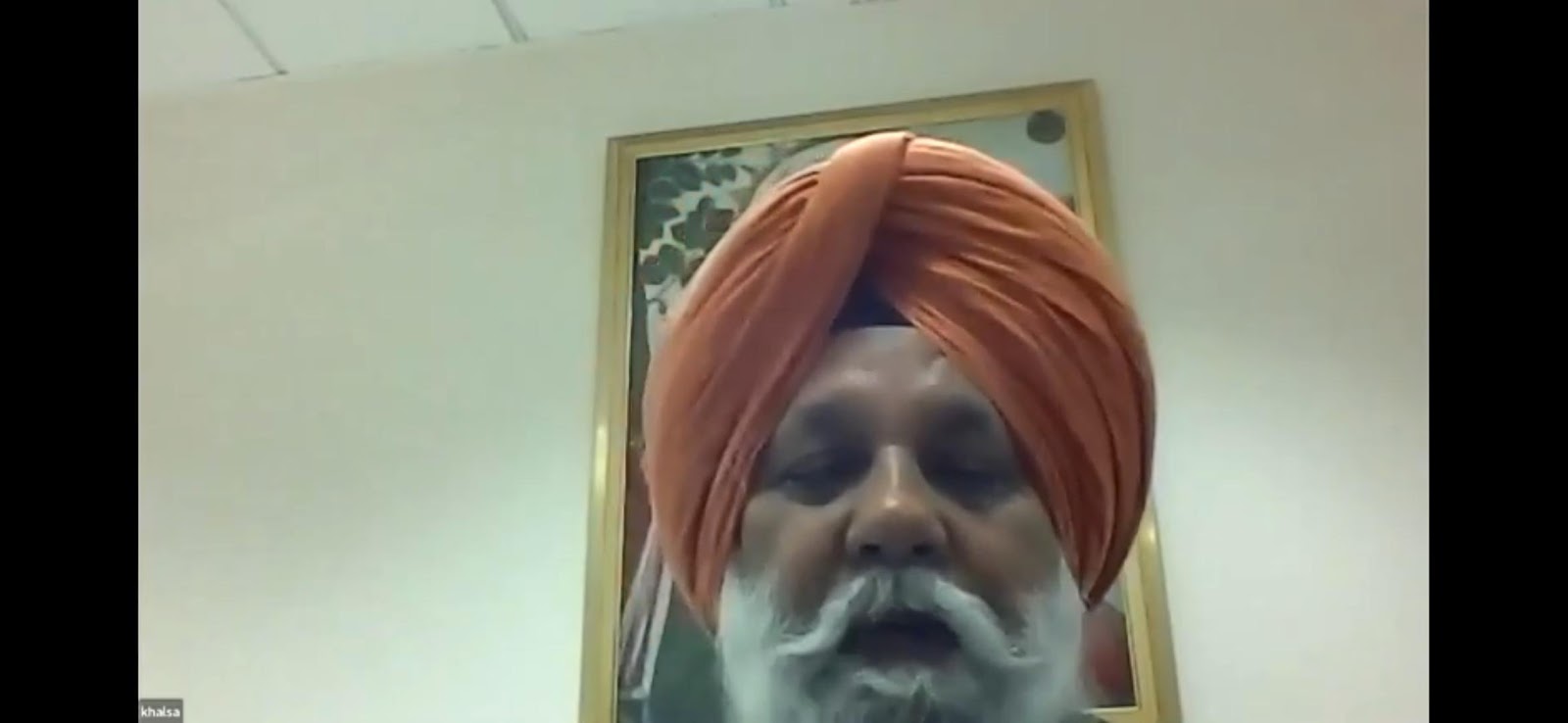 A close-up video still of a man with an orange turban and a white beard, appearing to speak, with a framed picture visible in the background.
