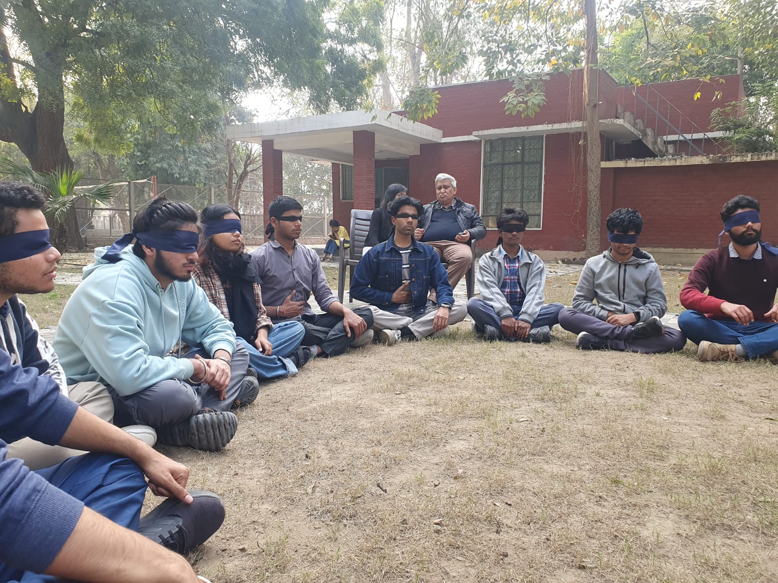 A group of young adults, mostly male, are seated in a circle on a patch of dry grass outdoors. Many of them are wearing dark blindfolds, while an older man watches from behind.