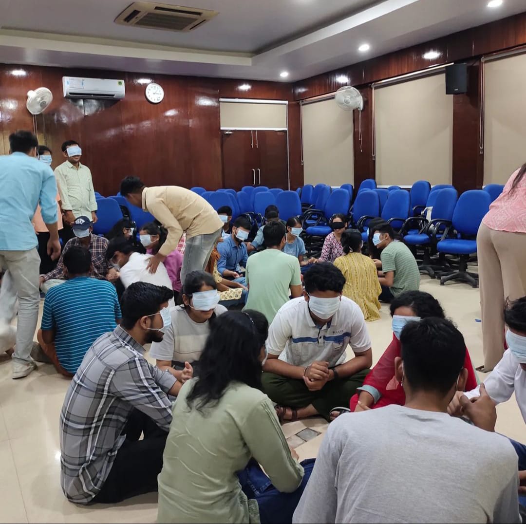 A diverse group of around 20 young adults are seated on the floor in a large room with wooden paneling and blue chairs stacked at the back. Many participants are wearing blindfolds or face masks, suggesting an interactive workshop focusing on sensory experiences or trust-building exercises.