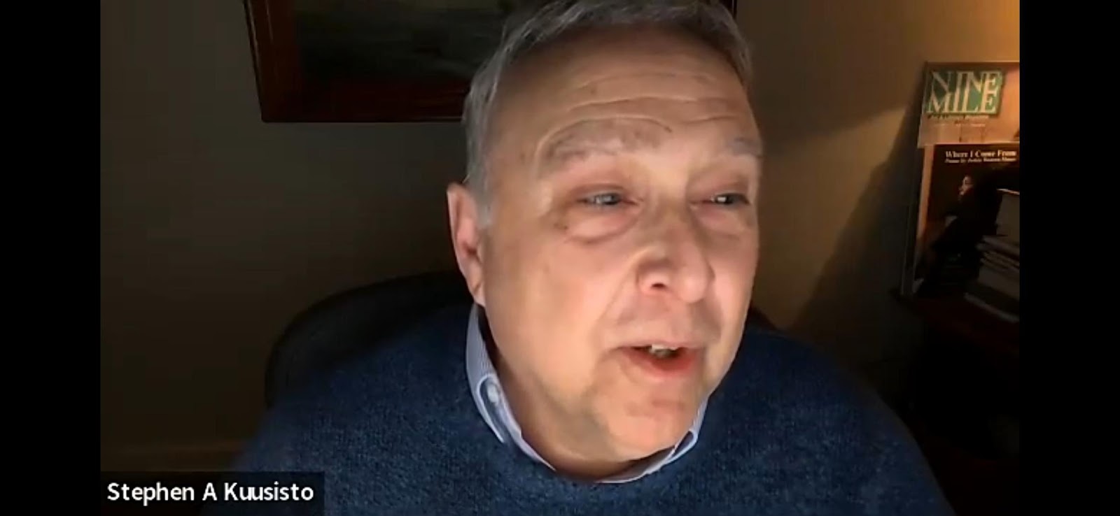 A close-up of Stephen A Kuusisto, an older man with grey hair and a blue sweater, speaking during a video call, with a framed picture and books visible behind him.