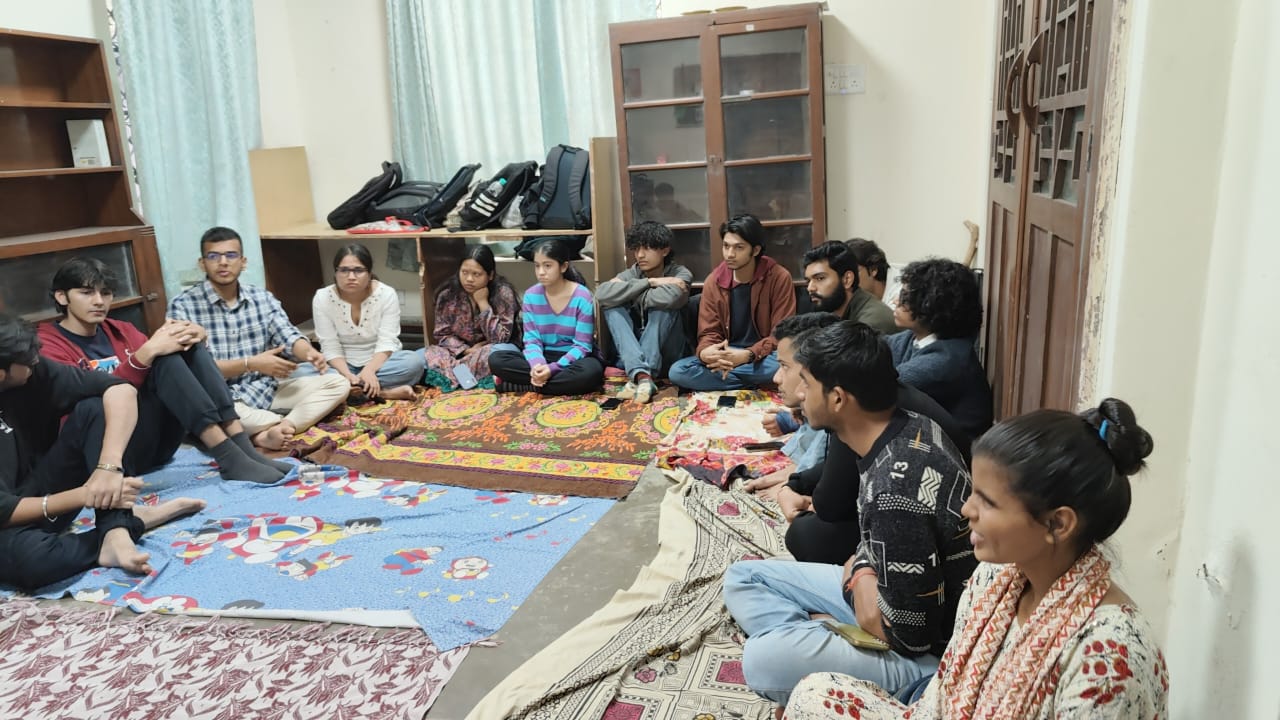 A group of diverse young adults are seated in a circle on patterned rugs in a casual room, listening attentively to someone out of frame. Bookshelves line the walls.