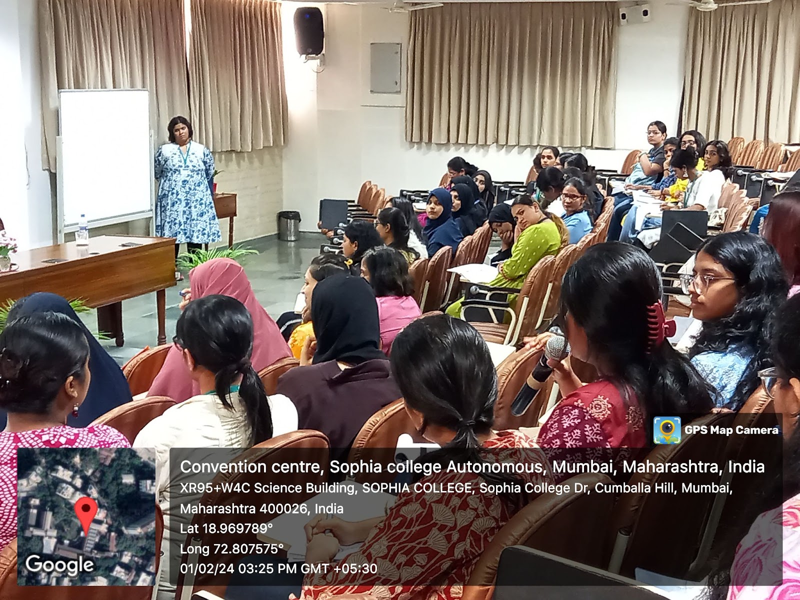 A female speaker in a blue and white patterned dress addresses a room full of young women seated in an auditorium, many taking notes, during a conference or lecture.