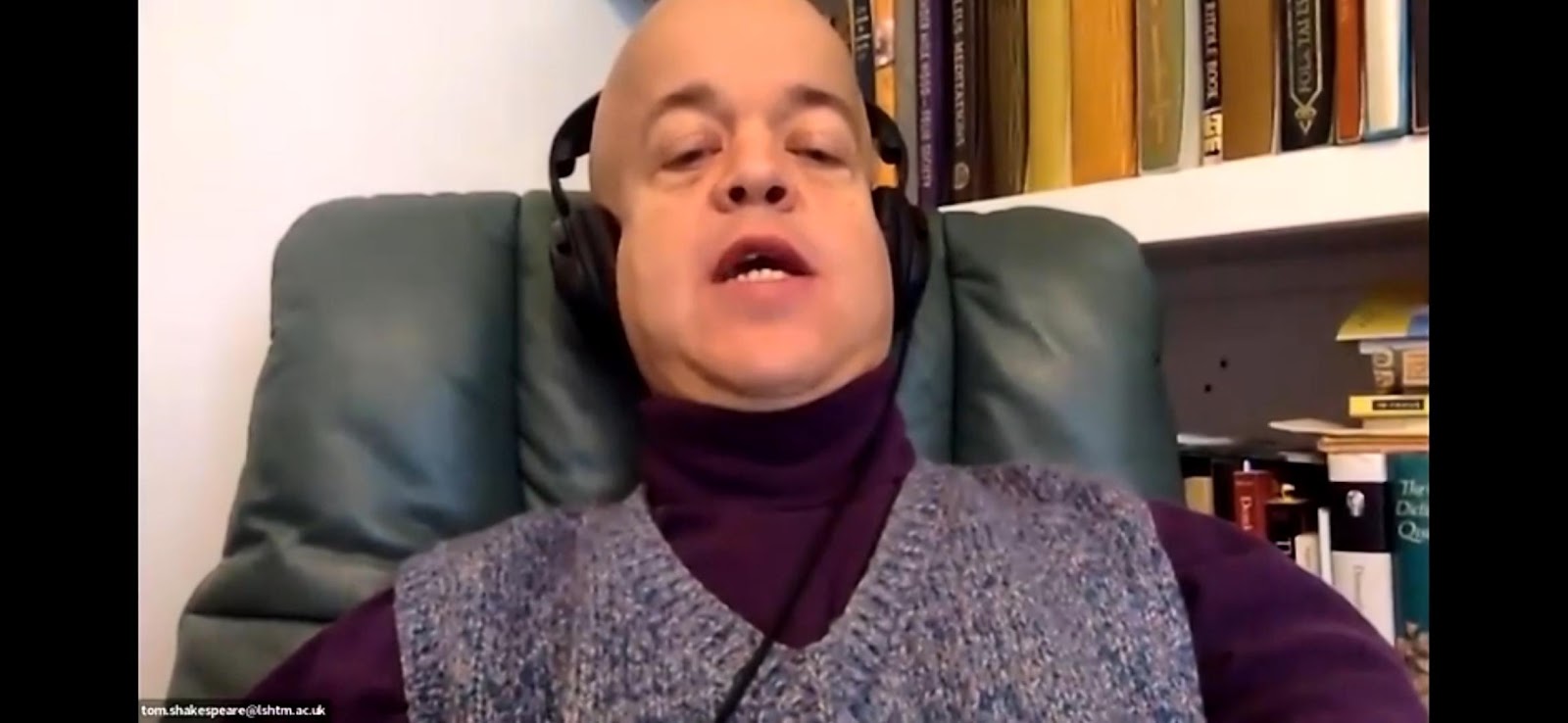 A man with a bald head, glasses, and headphones wearing a purple turtleneck and a patterned vest, speaking during a video call, with bookshelves in the background.