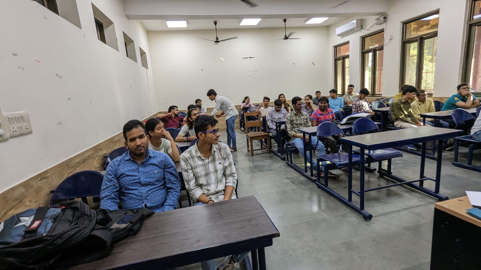 A wide shot of a classroom filled with around 20-25 students, mostly young adults, seated at desks. Some are looking towards the front, while others are interacting. The room has white walls, multiple windows on the right, and ceiling fans.