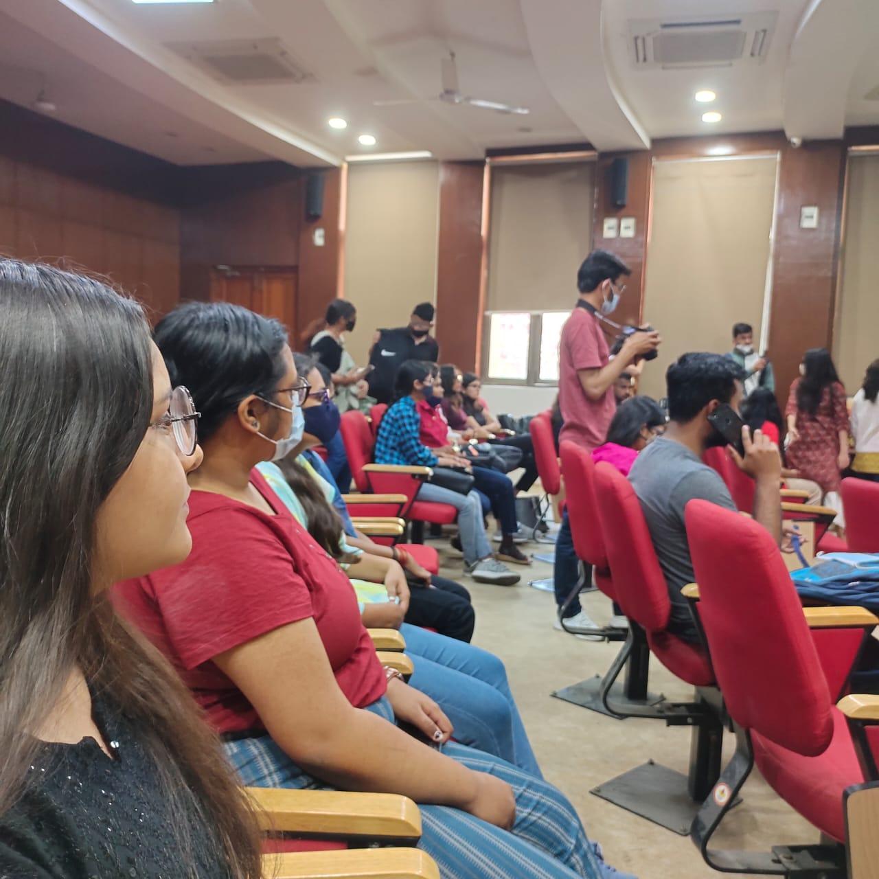 A low-angle, candid shot captures an audience seated in an auditorium, with a young woman in the foreground looking towards the right, and other attendees in the mid-ground.