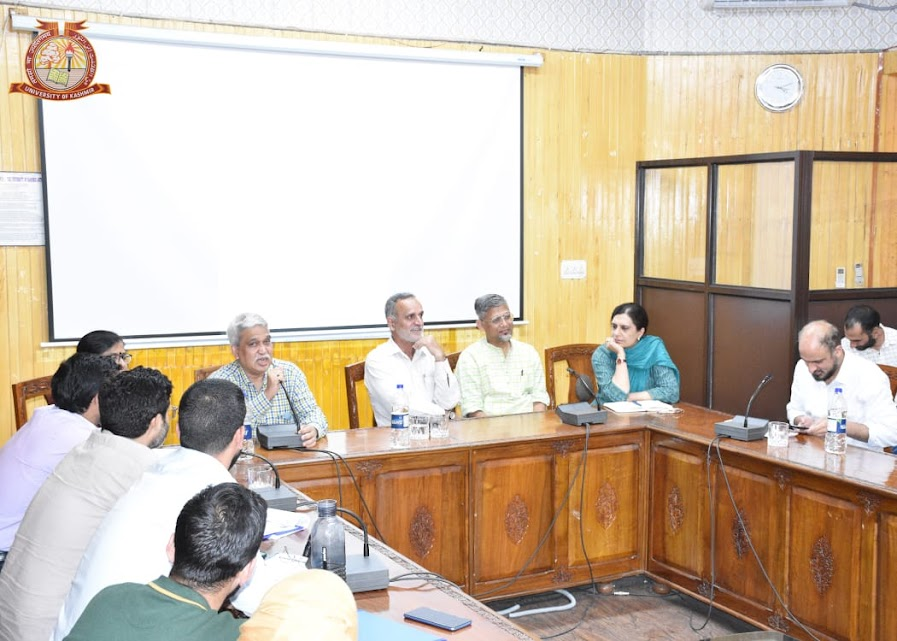 speakers and attendees sit around a conference table during a discussion session at the University of Kashmir, held as part of the Disability Studies workshop, with mics and notes in use.
