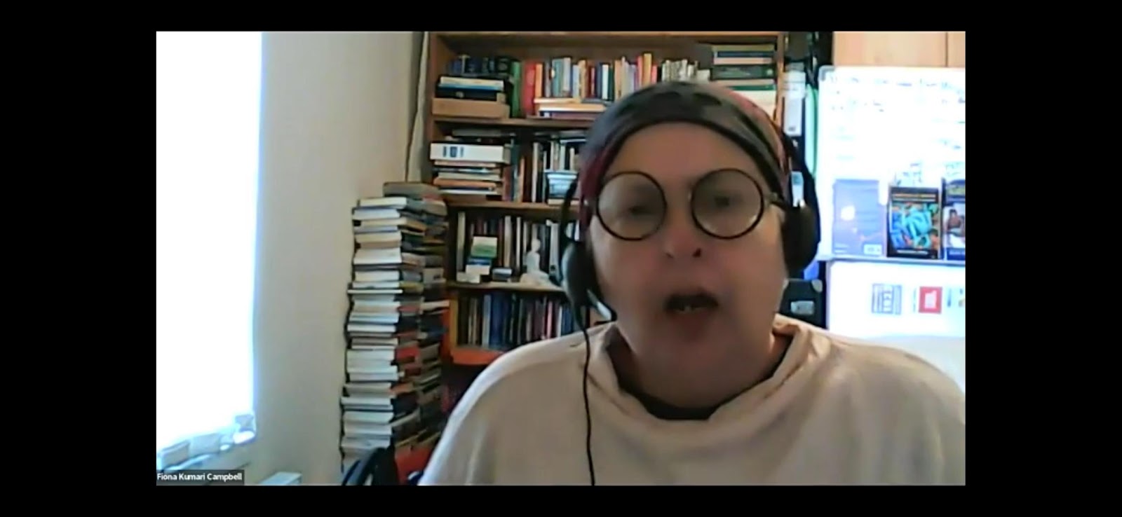 A person with round glasses, a patterned head covering, and headphones, speaking into a microphone during a video call, with bookshelves visible in the background.