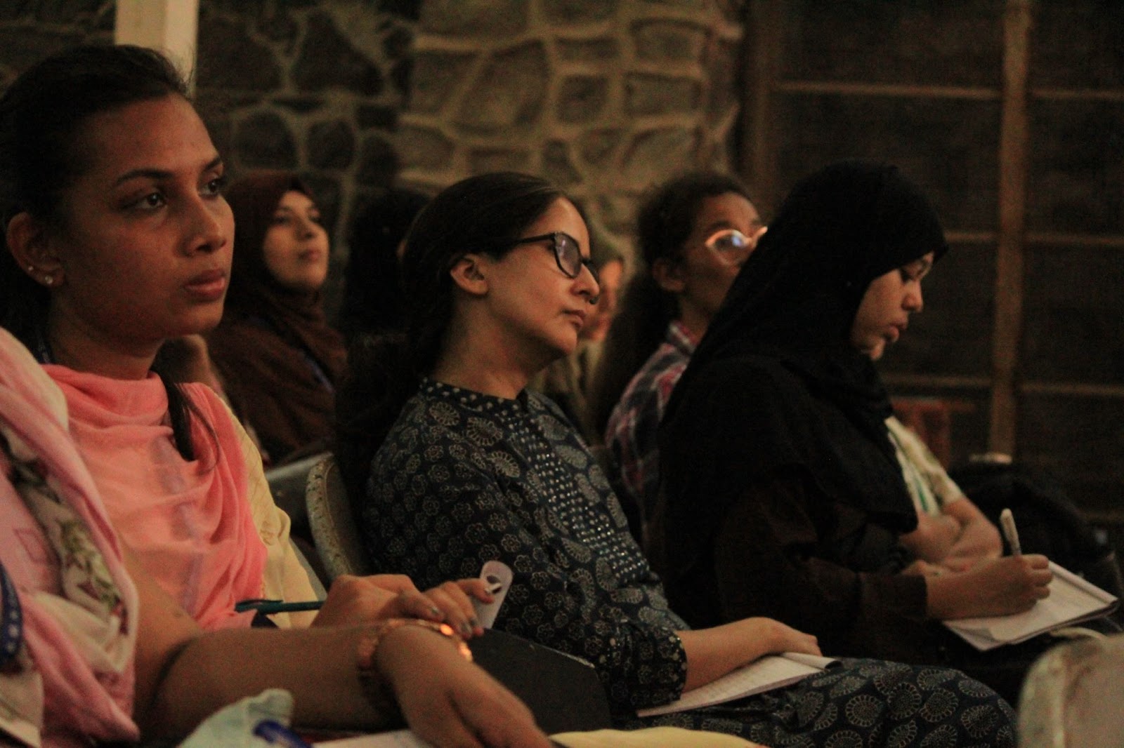 A row of diverse individuals, including a woman in a pink scarf, a woman in glasses, and a woman in a hijab taking notes, attentively listen in a dimly lit room with stone walls.