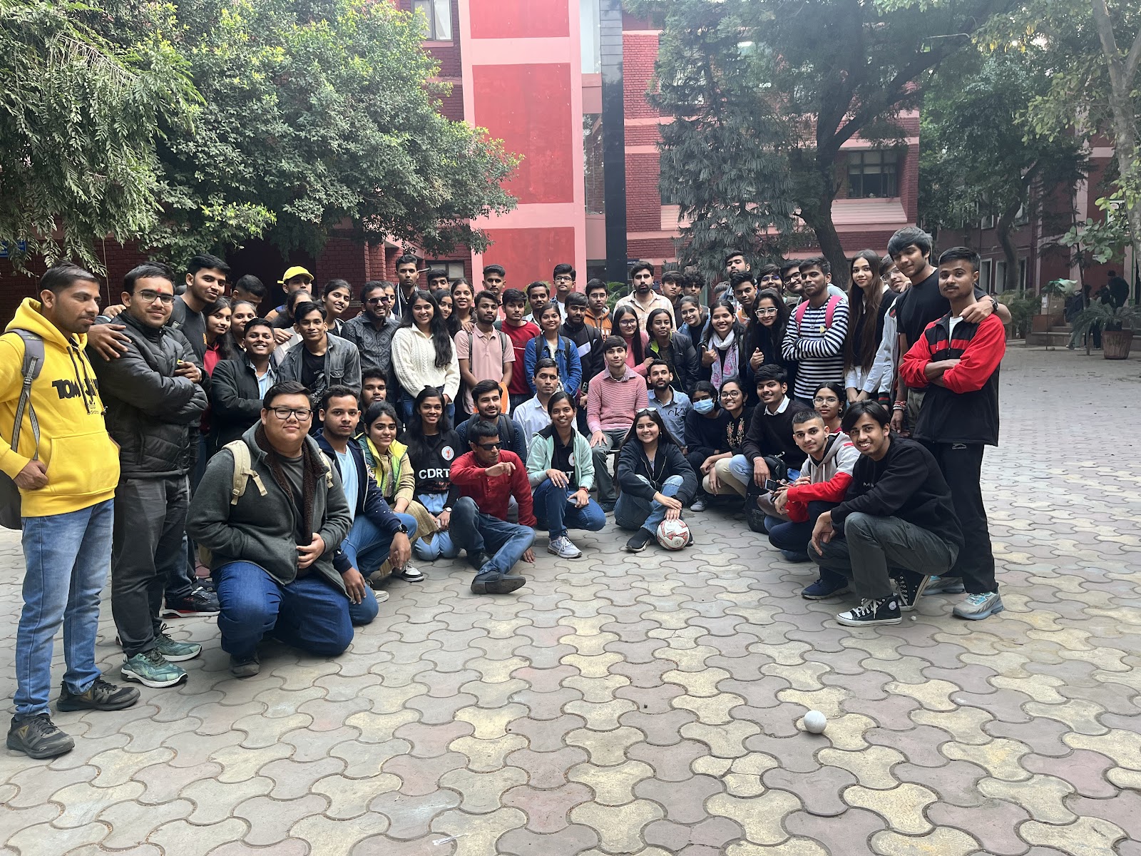 A large group of approximately 50-60 young adults, predominantly students, are gathered outdoors on a paved area with trees and a building in the background, posing for a group photo. 
