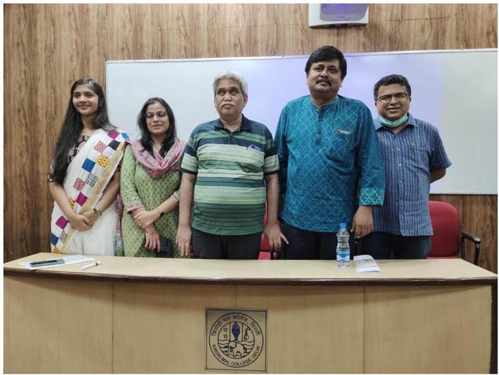 Five people, three men and two women, stand behind a wooden podium with a Kirori Mal College emblem, facing the camera and smiling, in a room with wood-paneled walls.