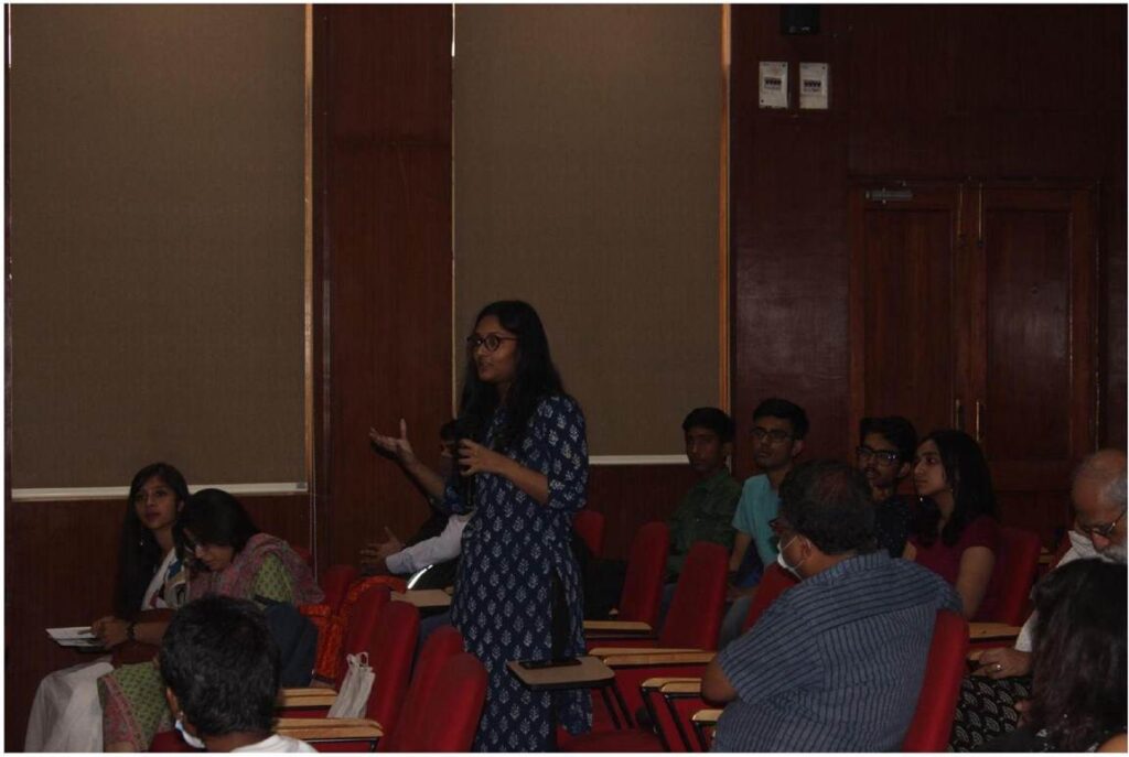 A young woman in glasses and a blue patterned kurta stands in an auditorium, gesturing as she speaks to a seated audience, with warm brown walls and red chairs.