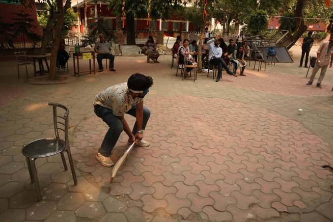 A person crouches, holding a bat, on a paved outdoor area. In the background, several people are seated on chairs under trees, observing the activity, suggesting a casual game or event in progress.