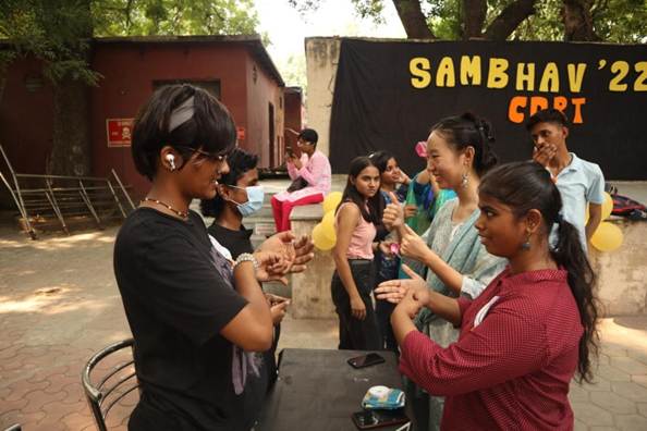 A lively outdoor event with "SAMBHAV '22" banner in the background. People are interacting, some appearing to be communicating via sign language, suggesting an inclusive gathering.