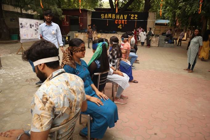 A group of people, some blindfolded, are seated outdoors, possibly participating in a sensitisation activity. A "SAMBHAV '22" banner is in the background, and other individuals observe the scene.