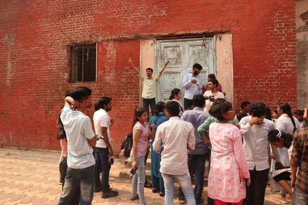 A diverse group of people gathers outdoors in front of an old red brick building with a faded double door. Some individuals are looking towards the door, while others are interacting amongst themselves.