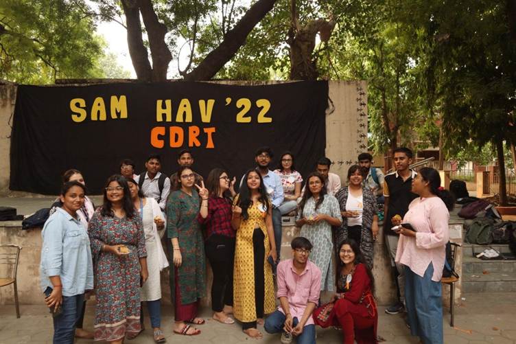A large group of young adults poses in front of a black banner with "SAMBHAV '22 CDRT" written in yellow. They are outdoors, surrounded by trees, appearing to be at an event.