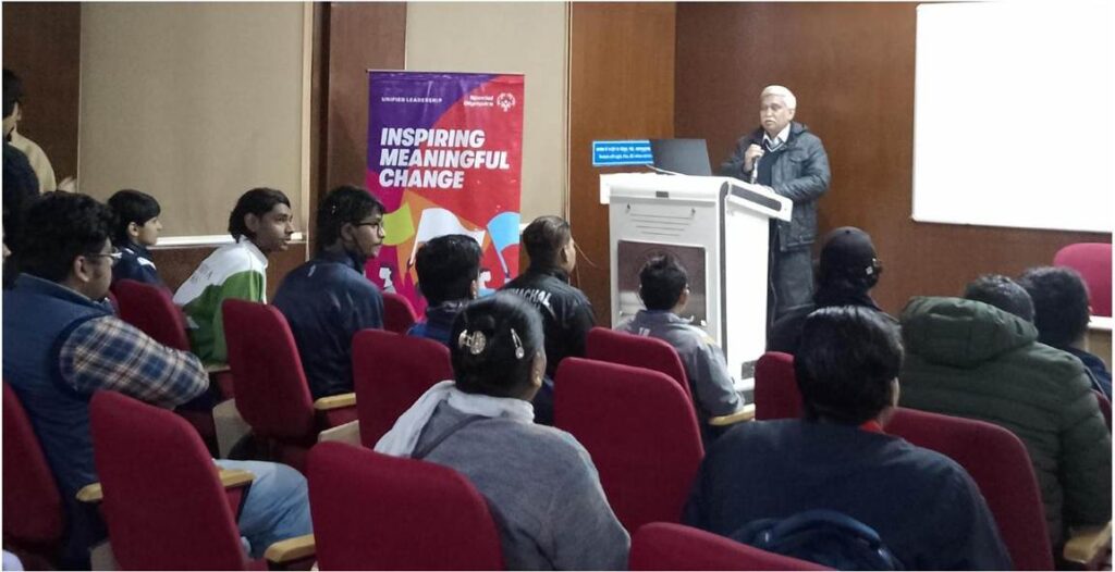 Professor someshwar sati in a dark jacket speaks from a podium to an audience seated in red chairs. A banner behind him reads "INSPIRING MEANINGFUL CHANGE."