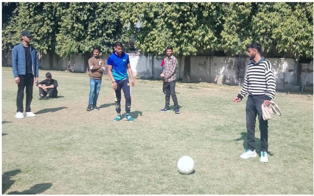 A group of six young men are on a grassy field, seemingly playing soccer. One man in a blue shirt stands near a white soccer ball, while others watch.