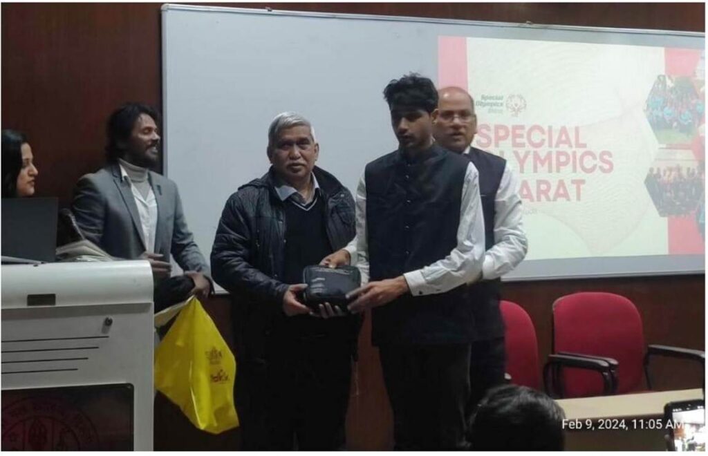 A young man in a black vest receives an award or gift from an older man in a black jacket, with a "Special Olympics Bharat" screen visible in the background.